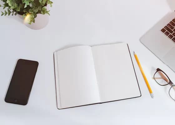 Białe biurko z góry: telefon, otwarty pusty notes z ołówkiem, okulary i róg laptopa
