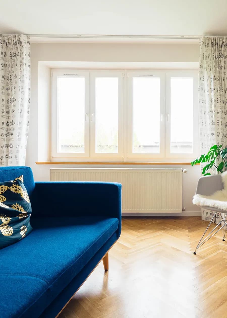 Salon w wynajmowanym mieszkaniu: niebieska sofa pod oknem, parkiet w jodełkę i biała komoda z telewizorem